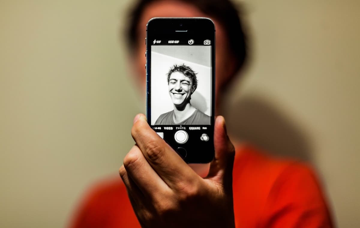 スマートフォンの画面にモノクロの笑顔の顔写真を表示し、背景に撮影された人物が立つ様子